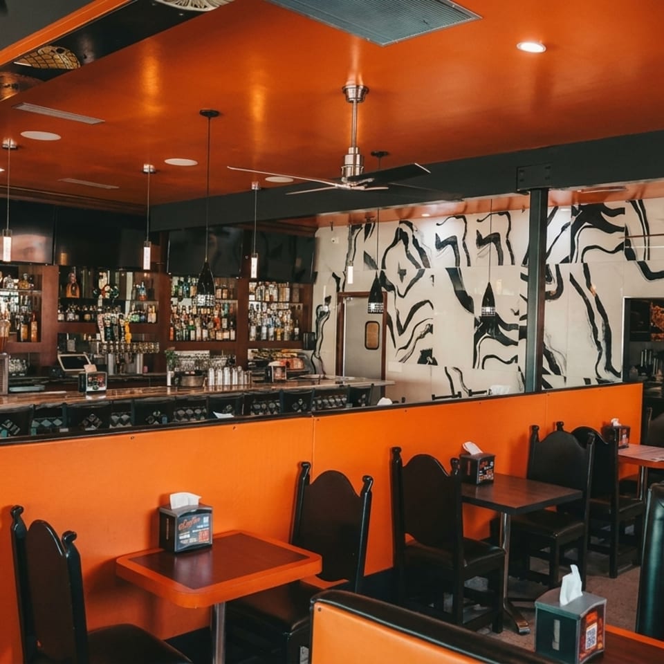 Vibrant restaurant interior with orange booths, dark tables, a full bar, and abstract wall art.